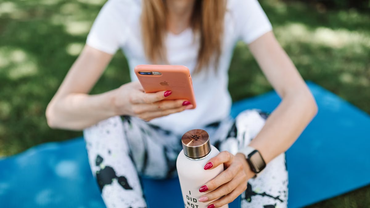 Woman using smartphone while holding bottle on yoga mat outdoors.